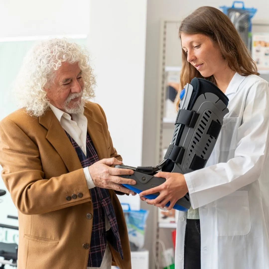 A healthcare professional in a white lab coat assists an elderly man with curly white hair as he examines an orthopedic walking boot in a medical supply store, with mobility aids and orthopedic products visible on shelves in the background.